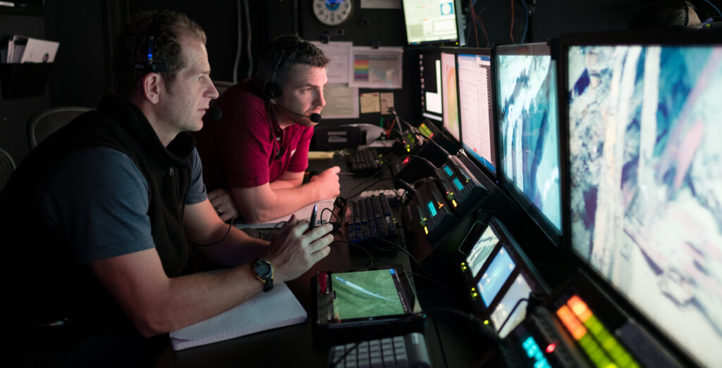 image of Dr. Daniel Wagner (left, JHT Inc./National Centers for Coastal Ocean Science) and Dr. Adam Skarke (right, Mississippi State University) in the control room of NOAA Ship Okeanos Explorer, 2018.