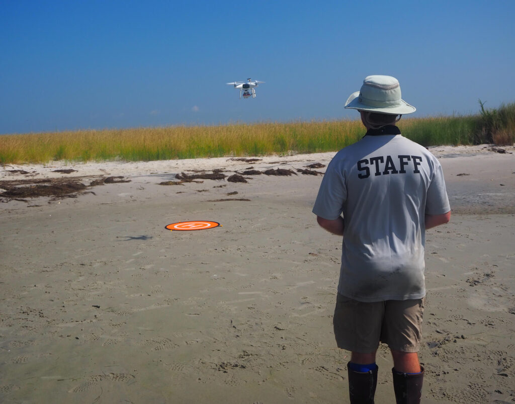 Person stands on a beach watching an uncrewed aircraft system hover near the dune.