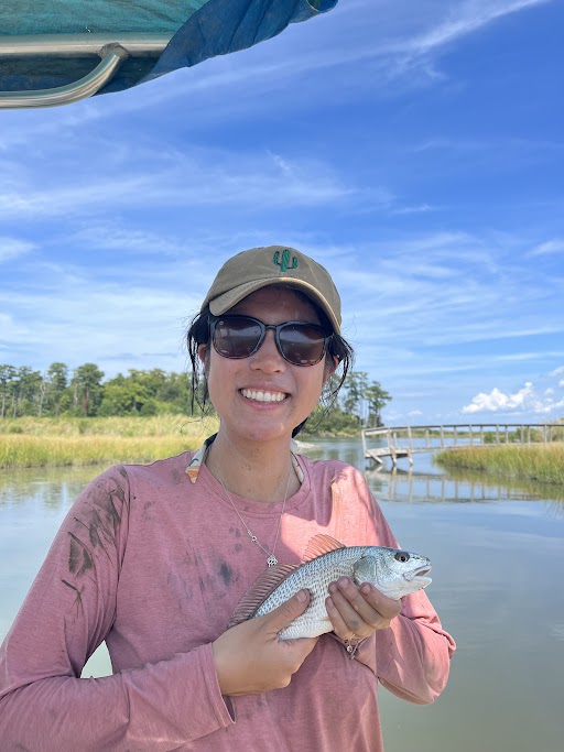 Claire Huang holding a fish with water, a pier, and marsh grass in the background.
