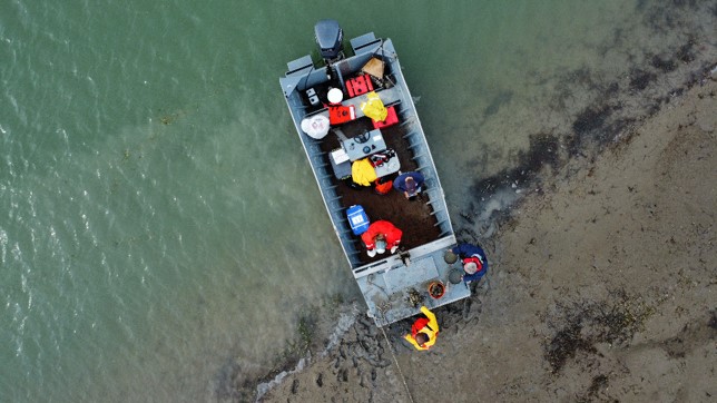 birds-eye view of a boat on a mudflat with two people standing on shore and one person in the boat
