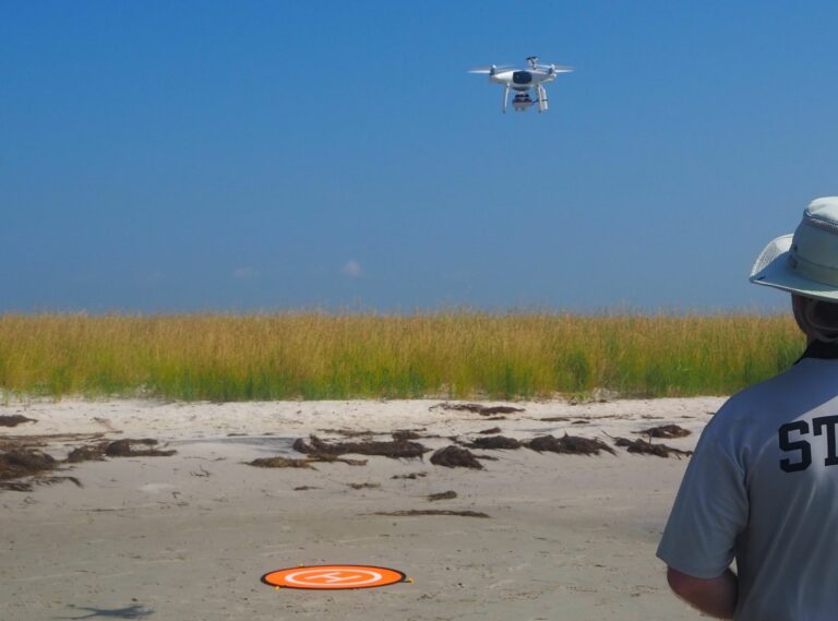 person on a beach faces a drone hovering over marsh grass