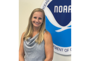 Blonde woman in grey dress poses in front of NOAA logo