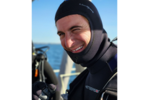 man wearing wetsuit hood smiles at camera