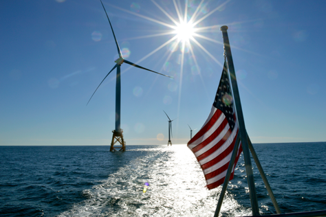 three wind turbines in the ocean on a bright sunny day, with an American flag visible in the foreground