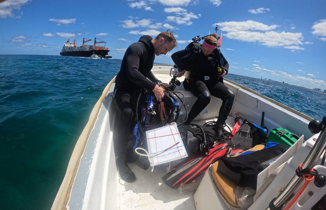 two scientists in SCUBA gear on a small boat on the water with a larger research vessel in the background