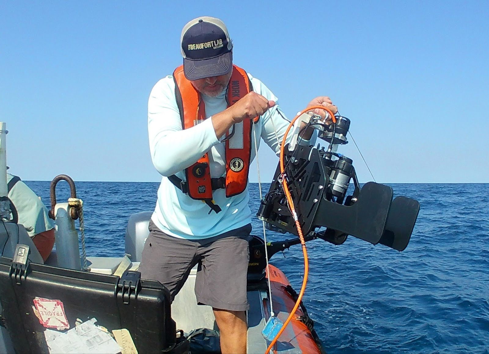 Scientist standing on a boat holds a large drop camera with water in the background