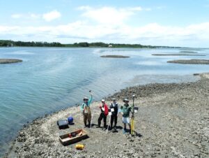 four people stand on an oyster reef with scientific equipment and wave to a drone. 