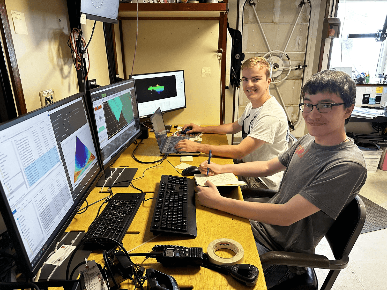 two young men sit in front of three computer screens showing seafloor mapping data 