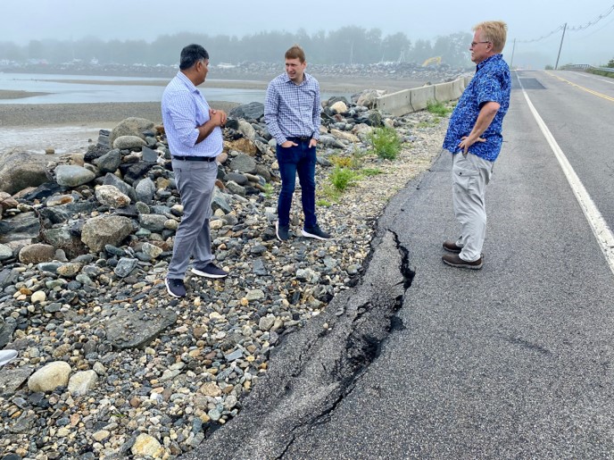 Three people stand along side a broken road lined with riprap