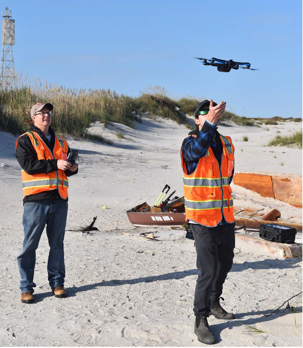 Two scientists are standing on a sandy beach. One is operating the remote control for an Uncrewed Aircraft System, or drone, and the other is about to catch the drone as it descends from flight.