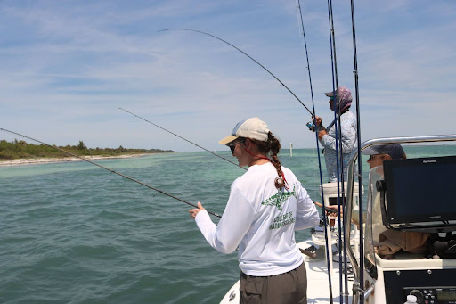 Three people stand on the side of a boat, holding fishing polls over the water facing the shoreline.