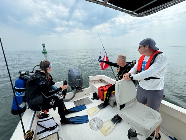 Three people in a boat. Two wear dive gear. One points at a buoy in the water. 