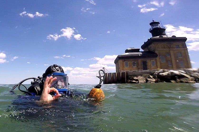 SCUBA diver at surface of water next to Toledo Harbor lighthouse gives "okay" gesture with right hand before descending to seafloor to collect mussels.